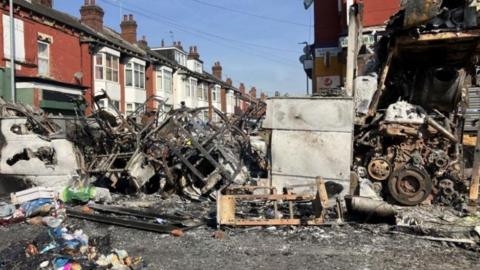 The burnt out shell of the bus that was torched during the disorder. The remains are also lying on the ground in front of a row of terrace houses.