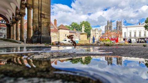 A square in York from which the minster and a red tourist bus are visible and reflected in a puddle