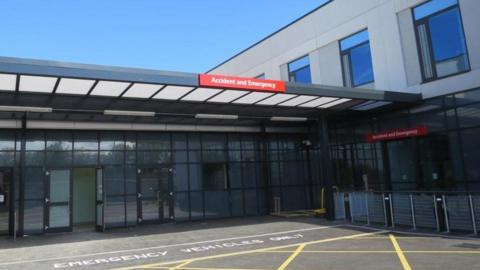 The entrance to Accident and Emergency at West Cumberland Hospital. It has a number of glass-fronted doors and windows and two signs with the name of the department above. It also has a parking area for emergency vehicles at the front.