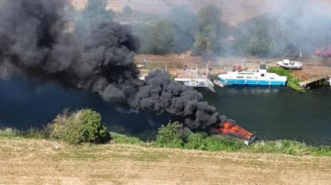 A photo taken from above by a drone shows thick black smoke billowing from a small burning boat in a dark river