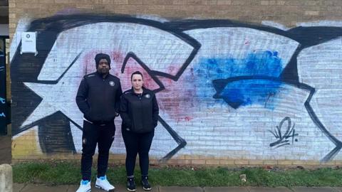 A man wearing a black jacket and trousers and white trainers. Standing next to him is a woman with a black jacket, black trousers and black trainers. They are standing in front of a white wall with coloured paint on it.