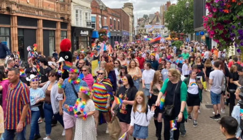 A large group of people making their way along Lincoln High Street. Some are brightly dressed in rainbow colours and some are holding flags. Other people are standing watching the parade.