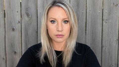 A woman with blonde hair in a black jumper and serious facial expression looking at the camera. She is standing in front of a grey garden fence.