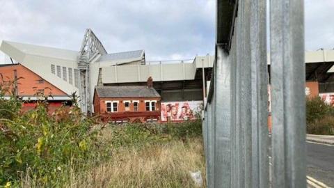 A patch of overgrown grassy wasteland with Anfield football stadium looming in the backdrop