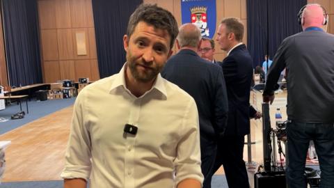 Man in white shirt stands in town hall in front of council banner.
