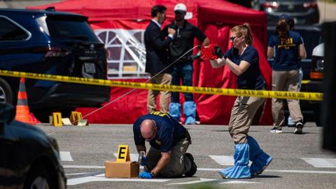 An image showing a pair of investigators behind caution tape surveying the scene of a shooting in Maine, where a 26-year-old man was shot and killed by an ICE agent. Behind them are an orange cone, a black van, other FBI investigators and a red tent.