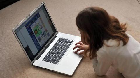 A girl with shoulder length brown hair lying on beige carpet looking at a laptop. 