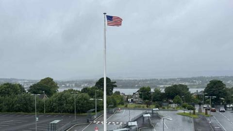 A US flag is pictured flowing in the wind on a large white flag pole.
