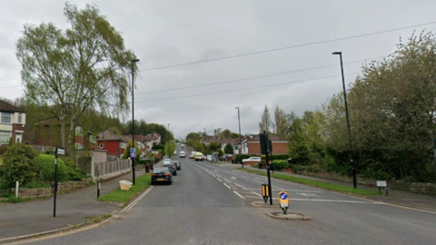 A suburban street with semi-detached houses to either side rises into the distance. Cars are parked on both sides of the road.