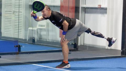 Andrew Simister plays padel on a padel court - a older man wearing sports clothes, a black top, grey shorts and trainers, leans over to take a shot. He holds a blue and green padel racket. He has a prosthetic leg.