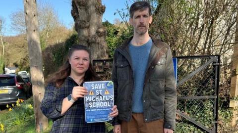 Claudia Parker and Geoff Black look at the camera. They are holding a sign which says 'Save St Michael's School.'