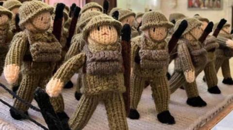 Four rows of knitted soldiers are marching in a scene. They are wearing green outfits and helmets, holding rifles to their left hand side 