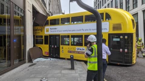 An image showing a yellow number 8 double-decker bus crashed into a stone and bronze metal shopfront, with smashed glass and a hubcap on the ground.