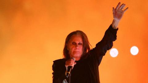 Osbourne with his left hand aloft performing on stage with microphone in his other hand. He is wearing a black T-shirt and blazer, heavy eyeliner and his dark hair is long.