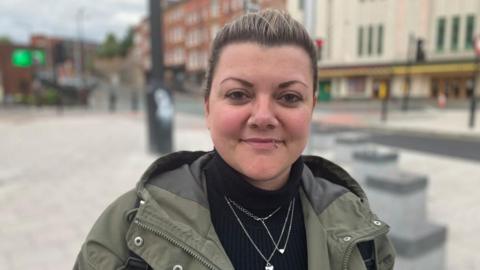 A woman wearing a camo jacket and black turtleneck stands smiling in Stockport's town centre. 
