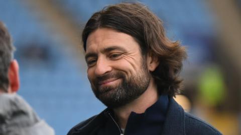 Russell Martin, wearing a navy blue jacket, smiles with blue seats blurred out of focus behind him at a football stadium