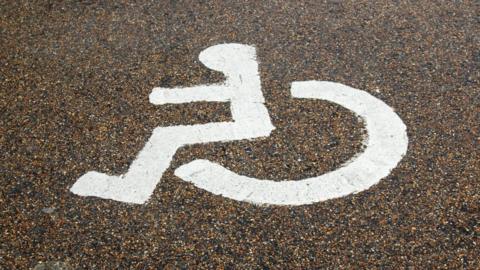 A white accessibility symbol on an asphalt surface. It is the familiar icon of a person seated in a wheelchair, typically used to mark disabled parking bays or accessible routes. 