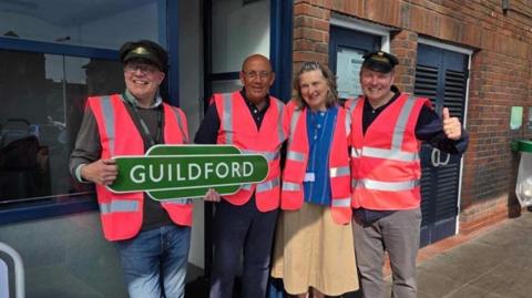 Train enthusiasts pose for a photograph while wearing bright pink, high-vis vests. A man on the left wearing a railway worker's cap holds a green sign reading 'Guildford'. They are stood in front of a brick wall at Guildford railway station.