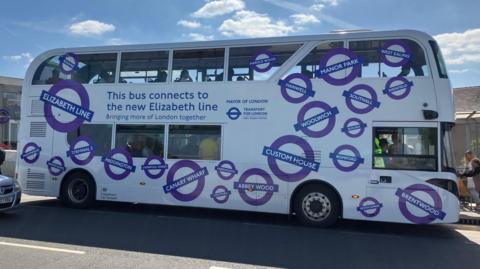 Double decker bus outside Abbey Wood station. There is a banner on the side that reads, "This bus connects to the new Elizabeth line". It is white and covered in images of Elizabeth line station roundels.
