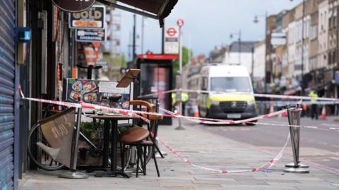 The exterior of a restaurant on a London street, the tape from a police cordon is wound around the crime scene, a chair is slightly tipped up beside a table. 