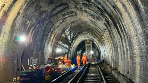 The Blackheath tunnel inspected by four men in orange jackets plus another using ladder. Equipment lays on the tracks to the left.