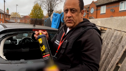 A man looking into the camera holding a nitrous oxide canister. There is a smashed car behind him.