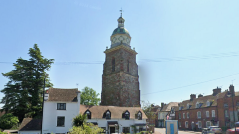 A white building with a thatched roof sits adjacent to a large brick tower. A dome structure sits at the top of the building, alongside a cross. Rows of houses can be seen surrounding the building.