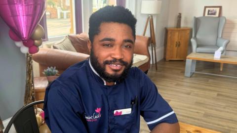 A man in a navy blue nurse uniform sits and smiles in a nursing home lounge area.