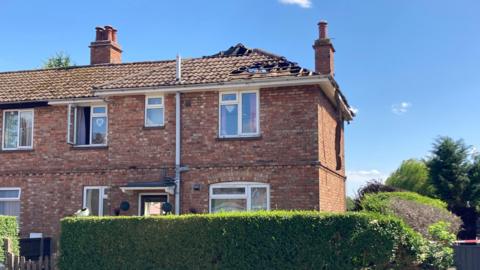 Brick house with a hole in the roof and two chimneys. There are five windows and a door plus two windows on the neighbouring property. There is a privet hedge in front of the house and trees to the side. It is a sunny day.