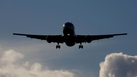 A Boeing 767 aircraft is silhouetted against a blue sky with fluffy white clouds. The aircraft is coming towards the camera with its wheels down ready to land.