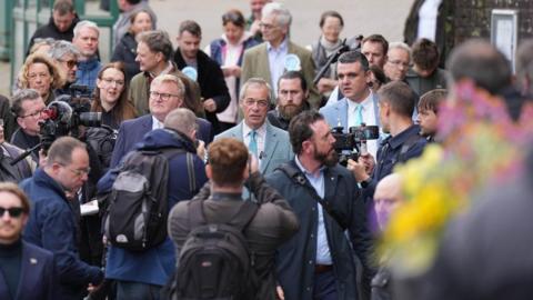 A group of people walk together along a street, surrounded by journalists and camera operators filming and taking photographs during a public visit.
