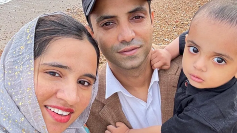 Nabeel Ahmad in a brown blazer, white shirt and black cap, holding his toddler son, who is wearing a black shirt and clinging at Nabeel, and Nabeel's wife on the left of the picture, wearing a hijab and has long hair tied back