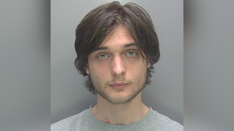 A custody photo of Jonathon Austin wearing a grey top. He is staring but not smiling. He has blue eyes, facial hair and long brown hair.
