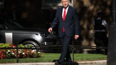John Healey walking down Downing Street wearing a suit with a red tie.