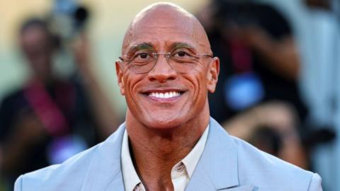 Dwayne Johnson smiles at the camera at The Smashing Machine red carpet event during last year's Venice International Film Festival. He is wearing an open-necked white shirt and a powder blue suit jacket.