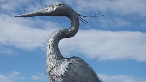 The head and neck of the giant heron, which is metallic in colour, with a bright blue sky with white clouds as a backdrop.