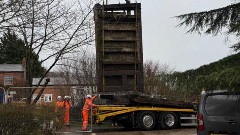 An old wooden lock gate being lowered by crane onto a yellow lorry trailer. A  worker in orange hi-vis and a white hard hat is guiding the gate down onto the trailer. There are two similarly dressed people behind him. There is a grey van to the right of the picture and canalside houses to the left.