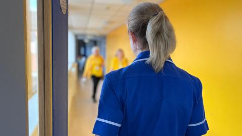 Over-the-shoulder view of a blonde-haired midwife wearing a blue uniform with white trim walking along a yellow-walled corridor with two people walking towards her