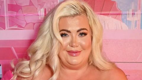 Gemma Collins is photographed looking into the camera at an event. She has dark brown eyes, long eyelashes and long, bleached blonde hair. The background wall behind her is different shades of pink.