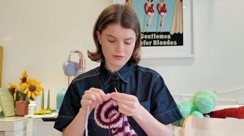 A young woman with shoulder length dark hair is knitting a pink stripped scarf. She is sitting in a bedroom with a bag of wool.