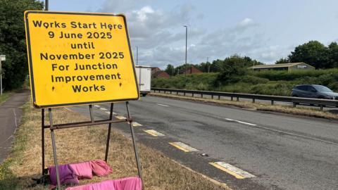 A yellow roadworks sign with 'work starts here 9 June 2025 until November 2025 for junction improvement works'. 
The sign is being held in position by a number of pink sand bags at the base. The sign is on the side of a dual carriageway. 