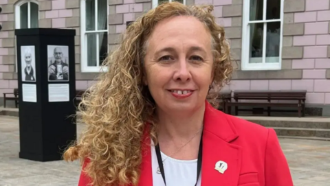 Deputy Inna Gardiner is wearing a red blazer and a white t-shirt. She has blonde curly hair and is stood in front of the States Assembly building.
