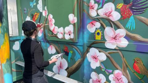 Katie O working on the Dosthill bus shelter for Dosthill primary school. The picture shows the back of an artist painting pink flowers and colourful birds on the shelter. the artist is wearing a black top, dark trousers and a black cap. 