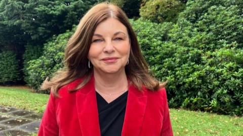 McLaughlin is smiling at the camera. She is wearing a red blazer and black top. she has shoulder length brown hair.