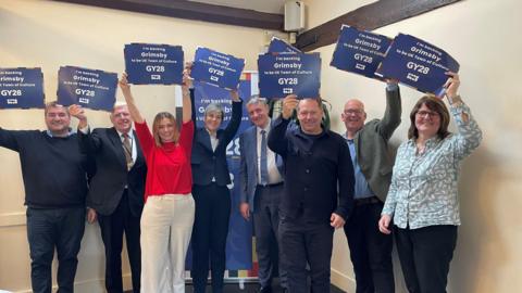 A group of people are smiling into the camera and holding up blue pieces of paper which say Grimsby GY28 on them. They are wearing work attire and standing in a room in front of a poster behind them.