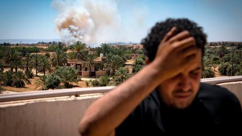 A Libyan fighter turns away as smoke rises in the distance