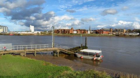 A small ferry boat is parked at a river jetty with the grassbank in the foreground and buildings lining the other side of the river in the distance
