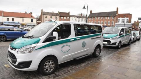 Several white taxis parked up in a taxi rank in Beverley town centre, they have East Riding of Yorkshire official green livery on the the doors and bonnet.