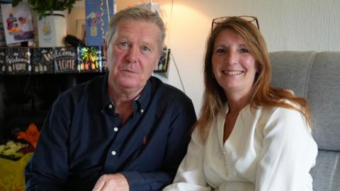 A man and woman sitting next to each other. She is to the right, smiling and sitting on a grey settee, wearing a cream blouse and has long red-brown hair. The man is to our left, crouching next to her and has a pensive expression and grey hair. He is wearing a dark blue shirt.