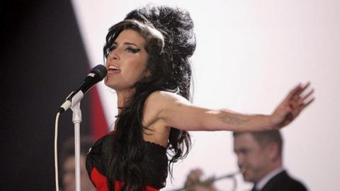 The singer with long dark hair in a messy beehive and black winged eyeliner sings into a microphone with her arms outstretched.